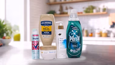 Five household and personal care products displayed on a kitchen counter: a bottle of Hellmann’s Real Mayonnaise, Degree Whole Body Deodorant spray, Dove Intensive Repair cream jar, Dove Pro-Ceramide body serum with pump, and Persil Wonder Wash laundry detergent in a green bottle. A blurred kitchen background with plants and utensils is visible.