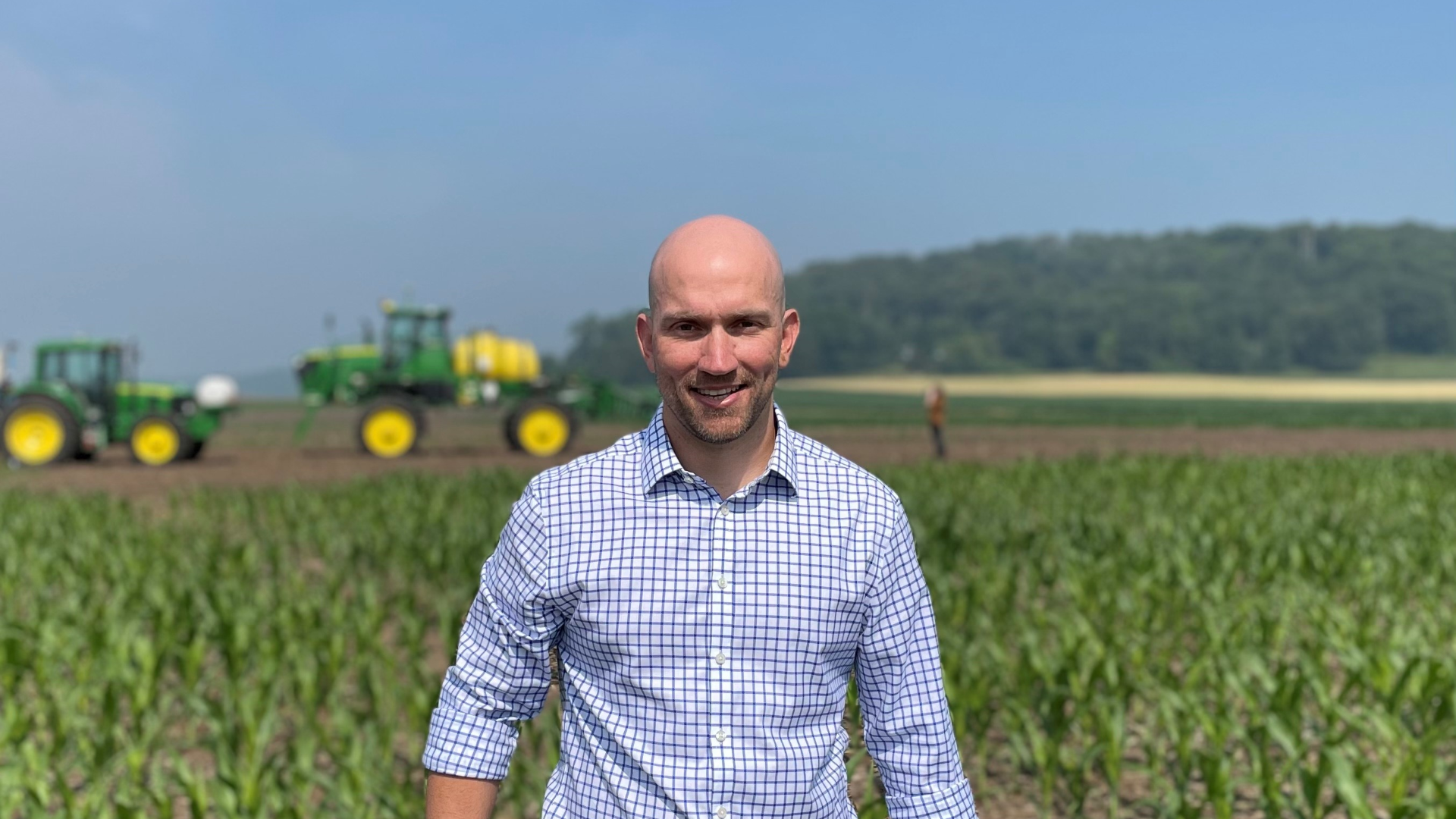 Lucas Urbano, wearing jeans and a checked shirt, stands in front of a field with two green and yellow tractors behind.