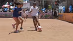 Girls playing football on an inner-city dirt pitch surrounded by fencing, being watched by a small group of people.