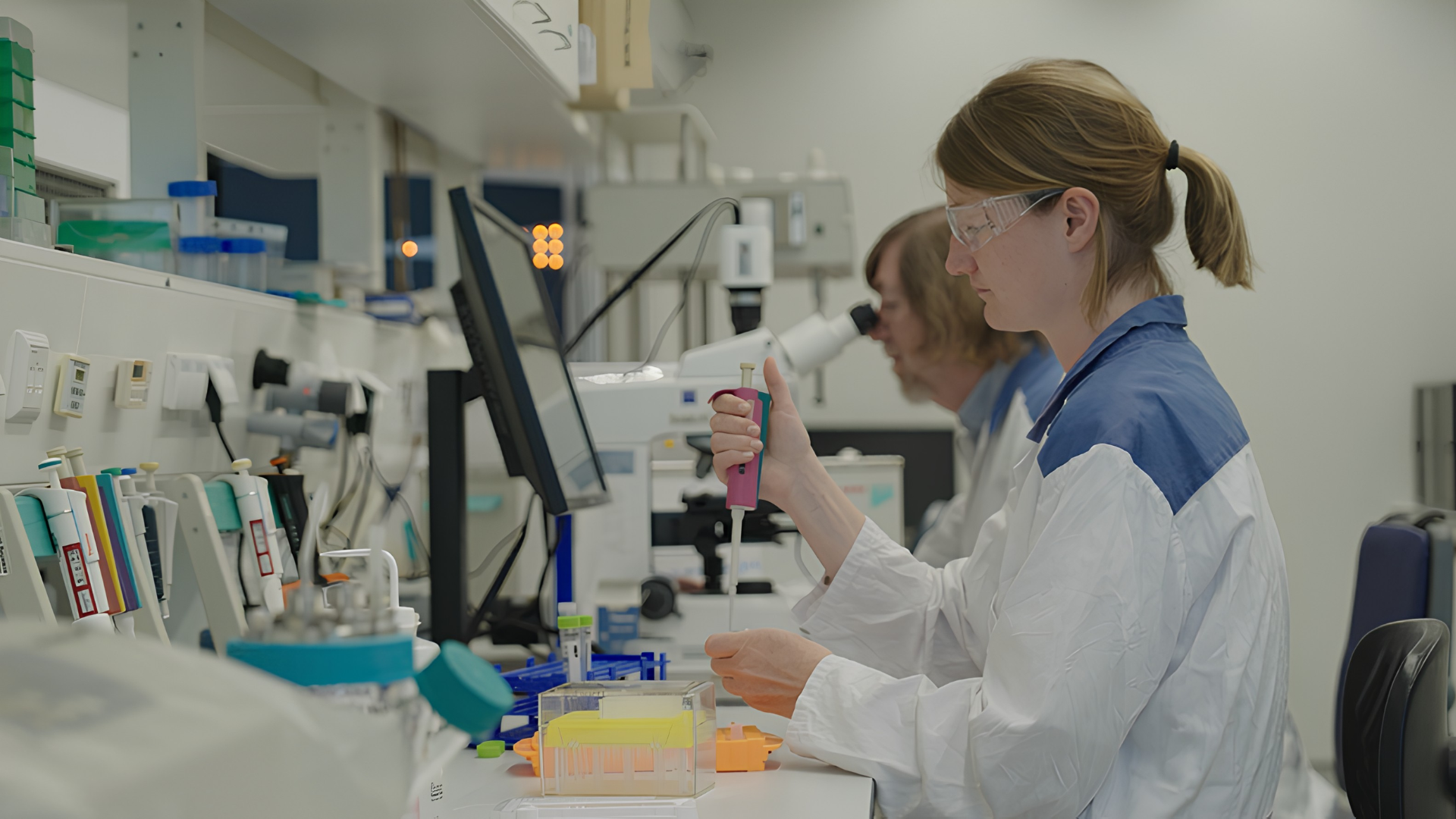 A woman in a lab coat and protective goggles uses a pipette.
