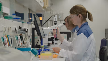A woman in a lab coat and protective goggles uses a pipette.