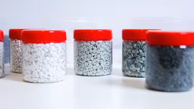Several transparent jars with red lids containing small plastic pellets in various shades, arranged on a white surface.