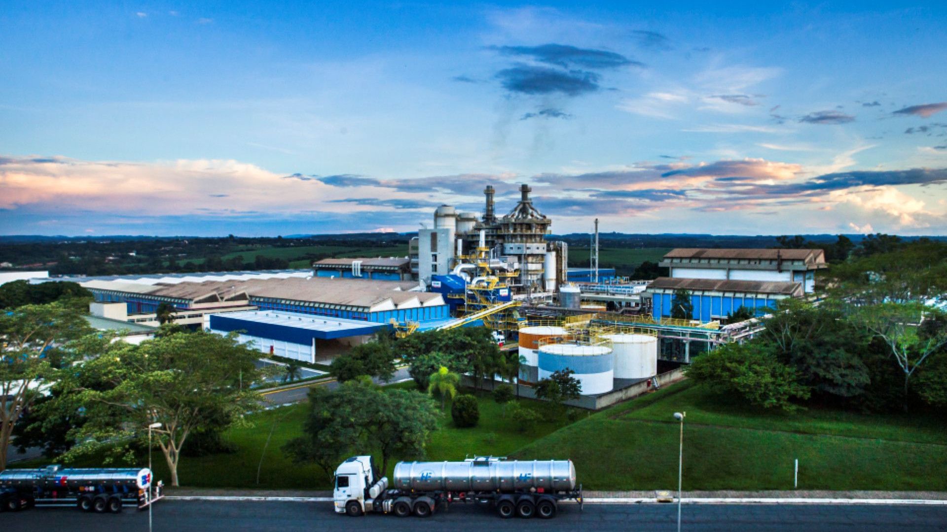Industrial site with tanks, buildings, and a tanker truck on a road, set against a blue sky with clouds and greenery.