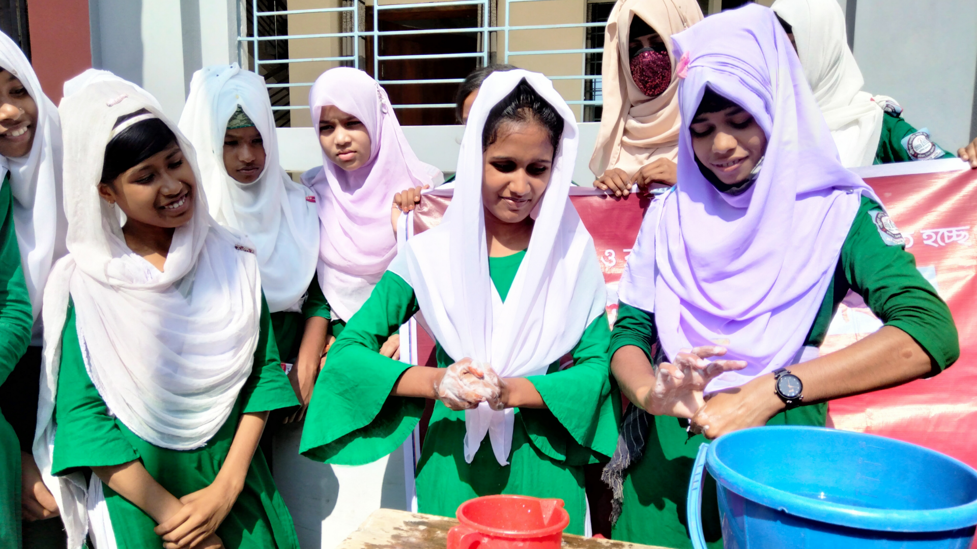 Schoolchildren in green uniforms with white hijabs washing their hands at a public tap