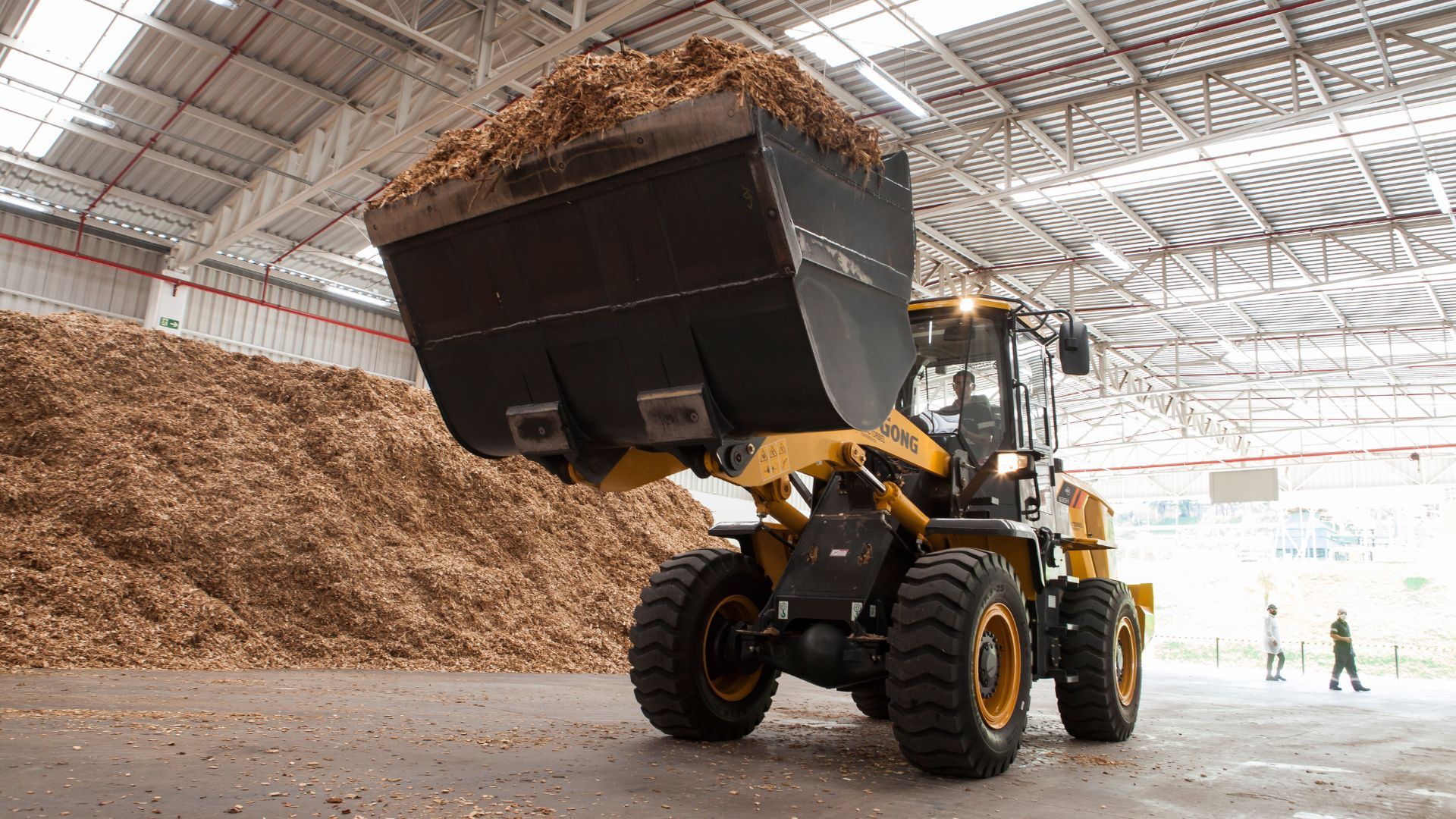 A tractor scoops up a front load of wood chips inside a large storage facility, with a large pile of wood chips and two people in the background.