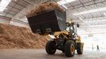 A tractor scoops up a front load of wood chips inside a large storage facility, with a large pile of wood chips and two people in the background.