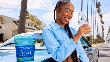 Woman in a blue shirt standing against a blue car, drinking a bottle of Liquid I.V.