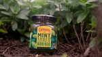 A jar of Colman’s mint sauce in front of green plants.