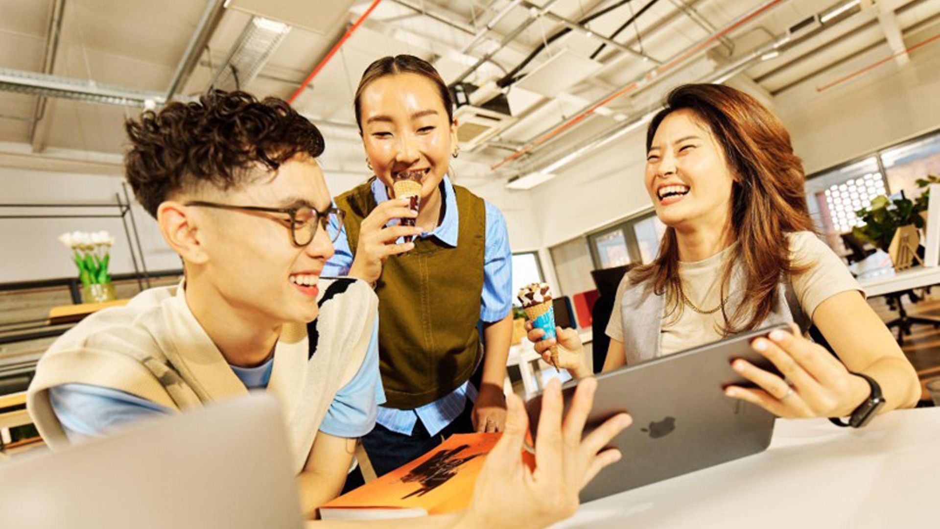 Three people in a modern office setting gathered around a table. One holds an iPad, and two of them are eating ice creams. Office furniture, plants, and shelves are visible in the background.