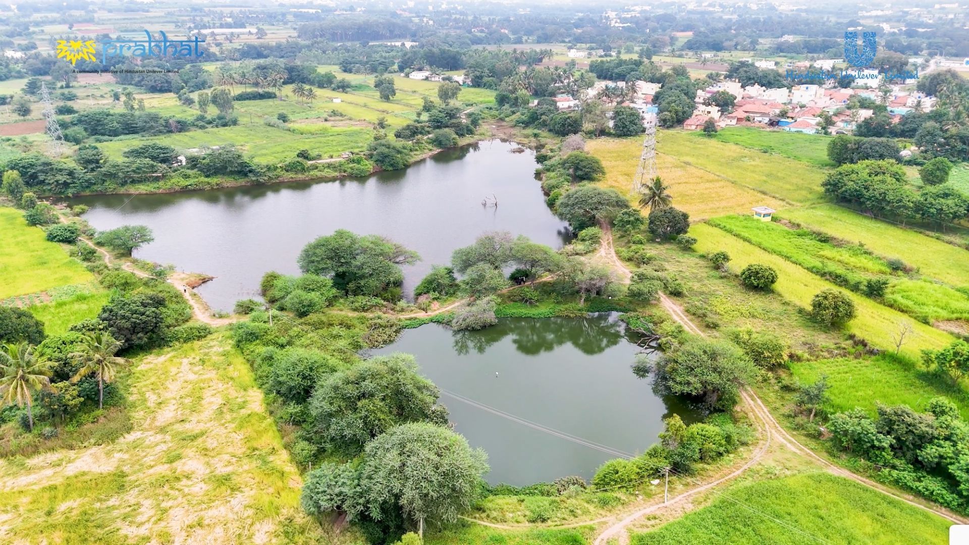 Aerial view of two small lakes surrounded by green fields, trees, and a nearby village settlement.