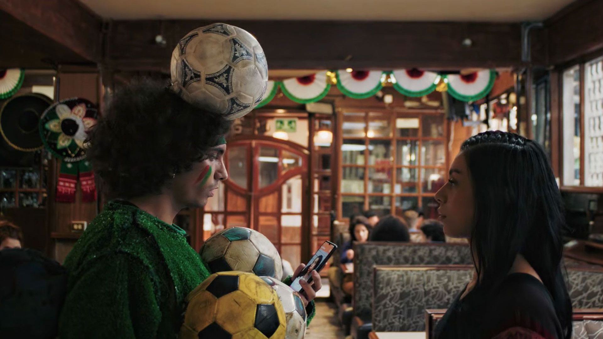 A person with a football balanced on their head and several footballs against their chest faces another person in a decorated pub with bunting and seated customers in the background.