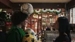 A person with a football balanced on their head and several footballs against their chest faces another person in a decorated pub with bunting and seated customers in the background.