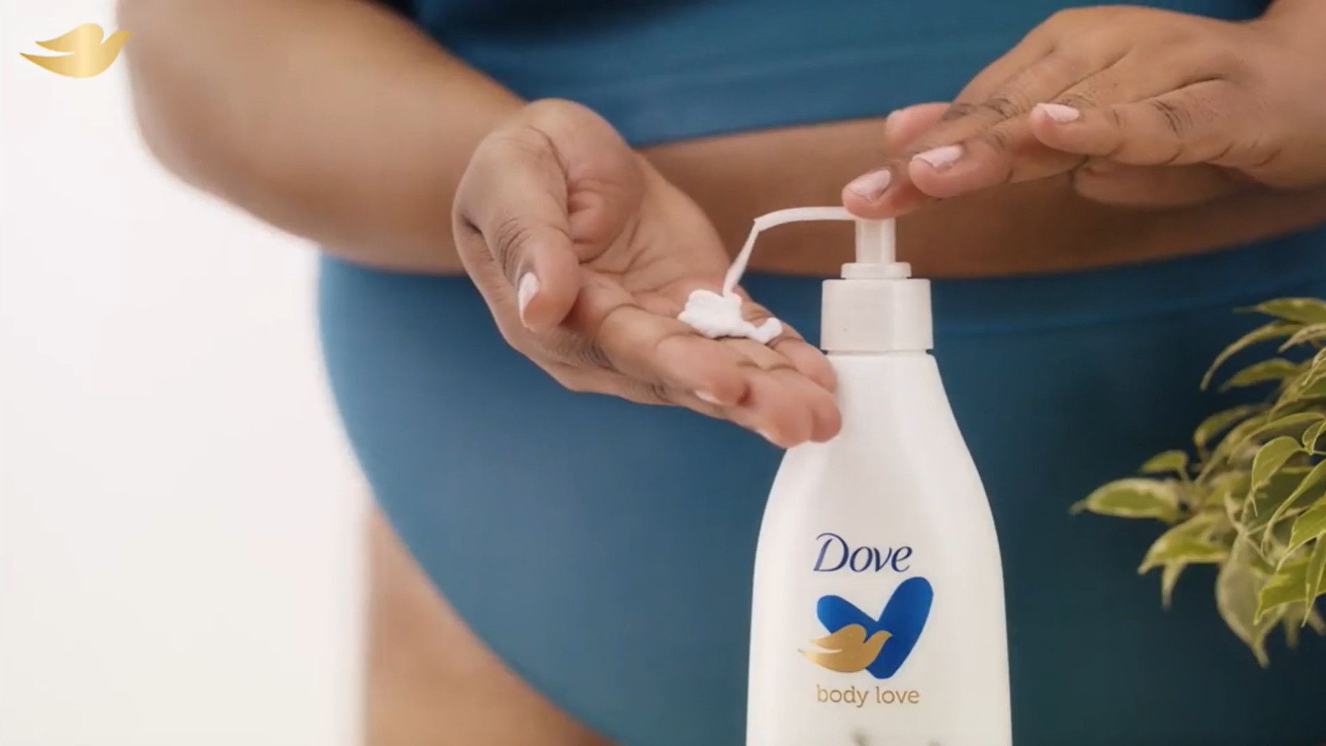 A woman in teal underwear pumps Dove Body Love lotion into her hand. Her face isn’t pictured. White bottle, Dove logo.