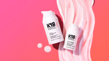 Two white bottles of K18 hair treatment placed on a pink background with swirled cream.