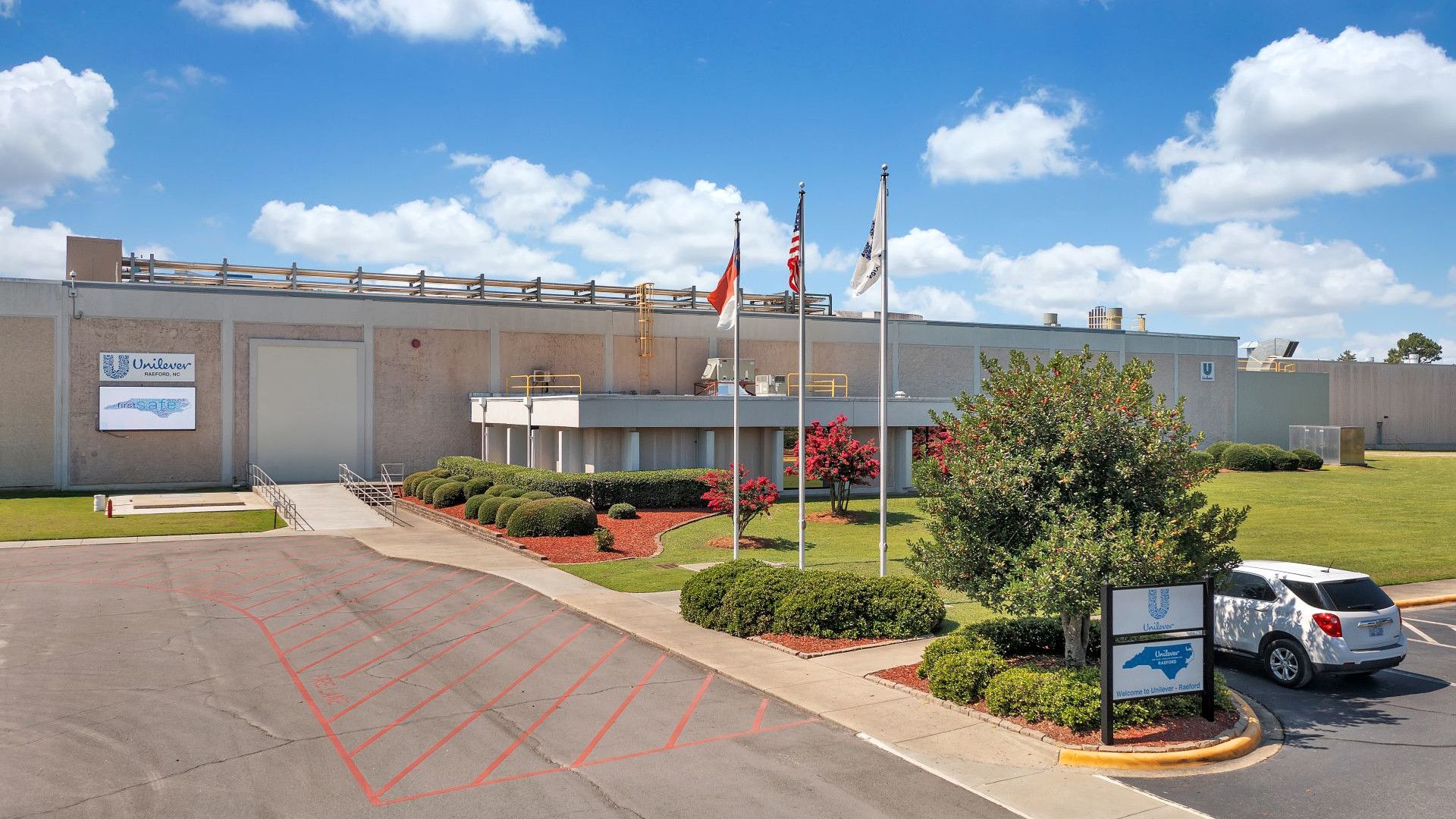 Front entrance of Unilever’s Raeford, USA facility with flagpoles, landscaped grounds and a parked vehicle under a bright blue sky.