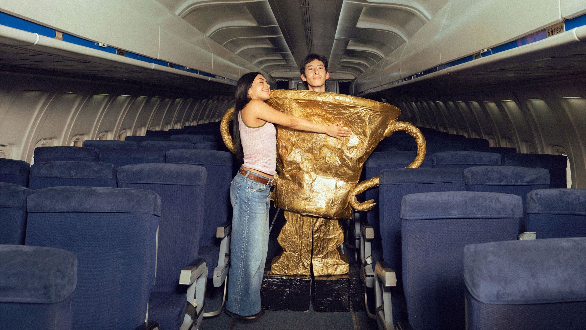 In the aisle of an empty aeroplane cabin with rows of blue seats, two people hold a large gold trophy prop resembling the FIFA World Cup. One person stands partly inside the oversized cup, with the rim around torso height, while the other stands beside it with both arms wrapped around the cup. The trophy has a crinkled metallic gold surface, two handles, and sits on a black base, with overhead luggage compartments visible along both sides.
