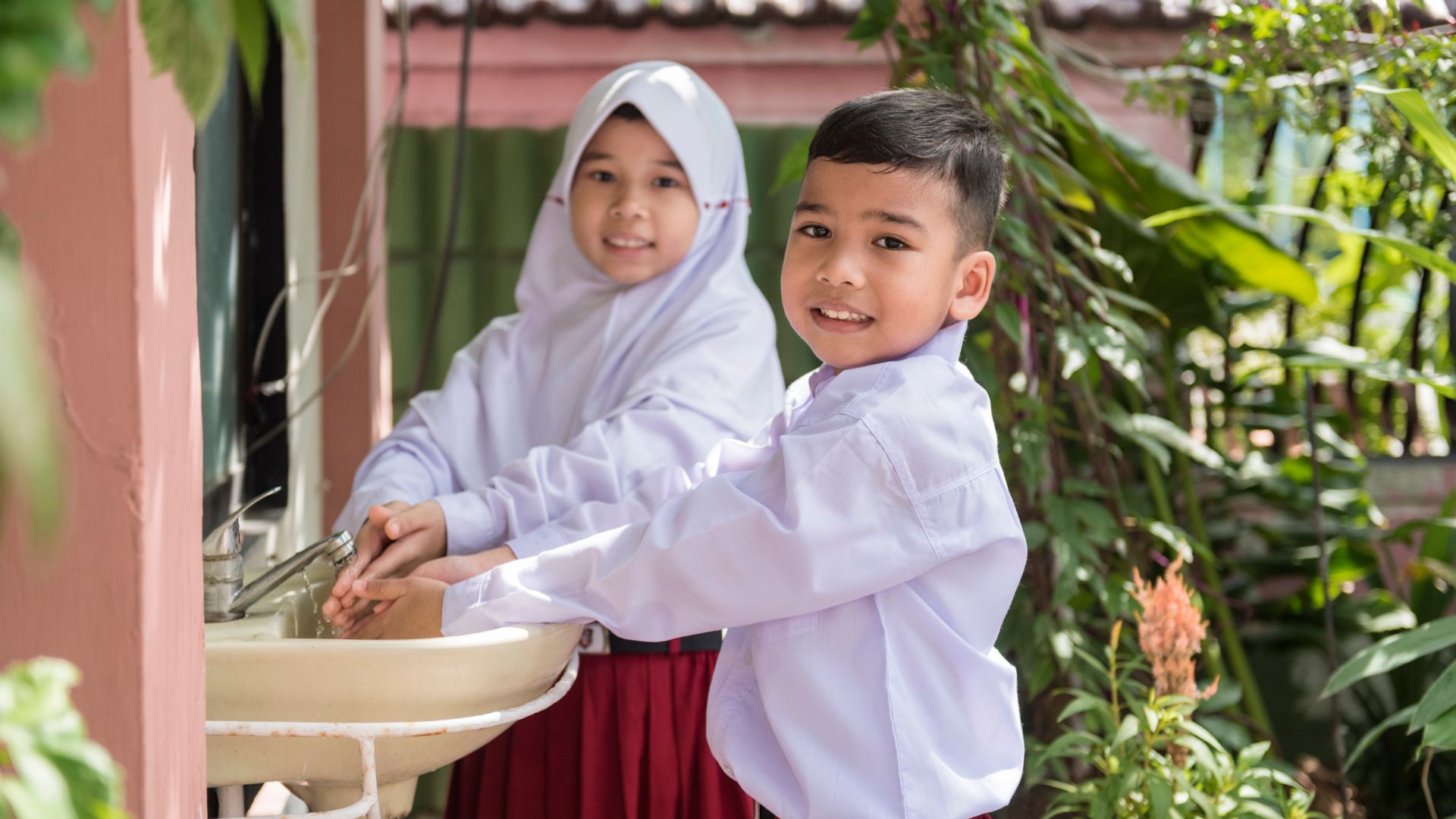 Two smiling Indonesian schoolchildren wash their hands at an outdoor sink.
