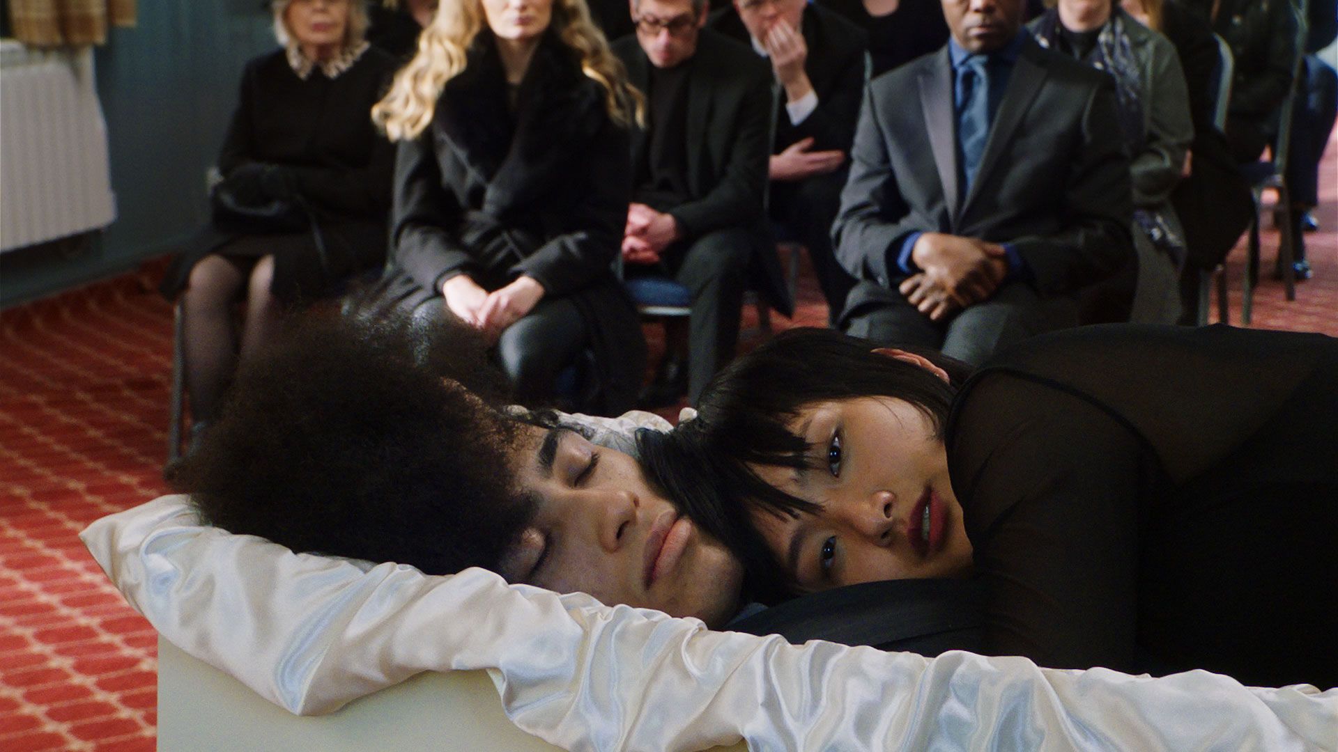 A funeral scene: a person lies in an open casket while a second person lies beside them inside the casket, looking toward the camera. Mourners in dark clothing sit in rows of chairs in the background.