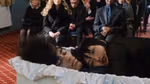 A funeral scene: a person lies in an open casket while a second person lies beside them inside the casket, looking toward the camera. Mourners in dark clothing sit in rows of chairs in the background.
