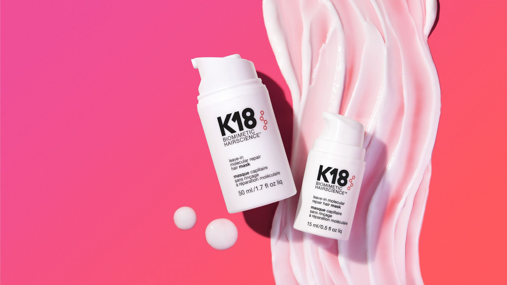 Two products from the K18 range – both white plastic bottles – lying on a bright pink background with a swirl of he product smeared on it.