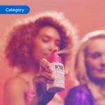 Hand holding a K18 hair product spray bottle with mist visible, set against a warm pink background.