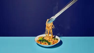 An image of Asian-inspired noodles being lifted from a white bowl with tongs, shot against a split background of dark and light blue.