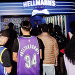 A crowd of people standing in front of a brightly lit Hellmann’s booth featuring a large illuminated Hellmann’s sign with a bowtie logo. The booth has a modern design with blue lighting, vertical light bars, and product imagery displayed along the walls. The scene appears to be part of a busy event or activation, with attendees gathered and looking toward the booth’s interior.