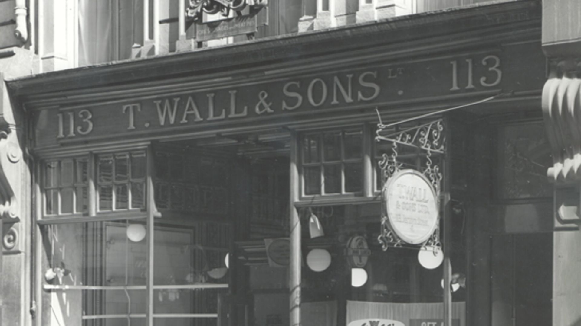 Black and white photograph of the storefront of T. Wall & Sons Ltd at number 113. The store name is displayed above the entrance, flanked by the number 113 on both sides. A hanging sign with partially legible text is visible in front of the store. Large display windows reveal various items inside.