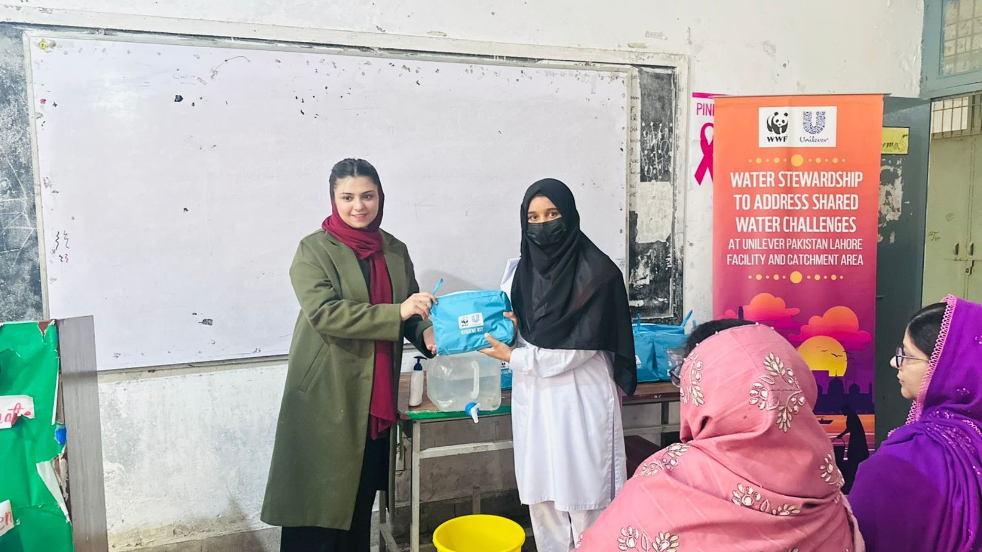 Two women at a community workshop hold a hygiene kit in front of a water stewardship banner.