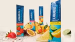 A variety of Liquid I.V. Hydration Multiplier electrolyte sachets in different flavours, including Strawberry and Lemon Lime. The packets are arranged around fresh fruits like strawberries, passion fruit, pineapple, lemon, lime, and orange slices on a light-coloured surface with water droplets.