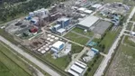 An aerial view of an industrial facility featuring buildings, large storage tanks and some green spaces, surrounded by roads and trees in the background.