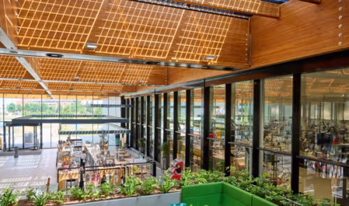 The interior of a bright and airy office building, with a foyer to the left featuring a canteen and various plants. To the right, behind glass panels, is a large office space.