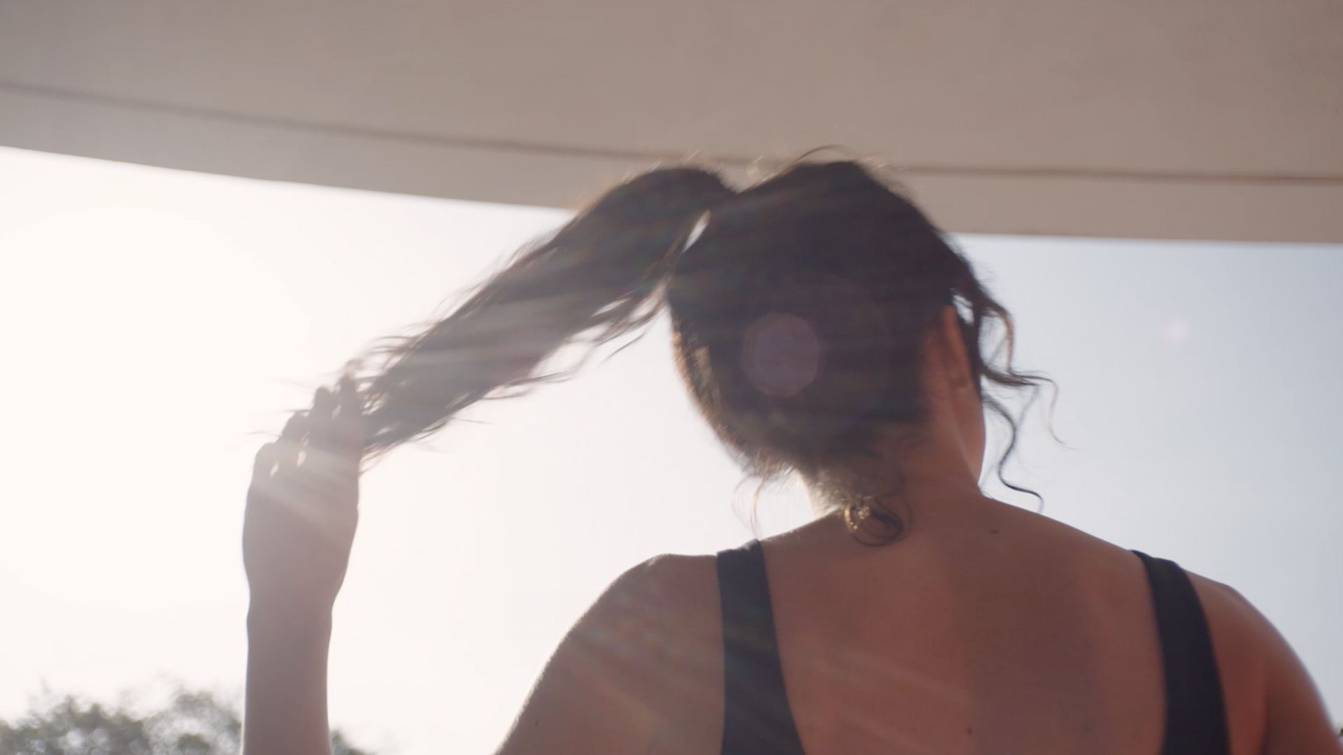 Back view of a person holding their ponytail in bright sunlight with lens flare, wearing a sleeveless top.