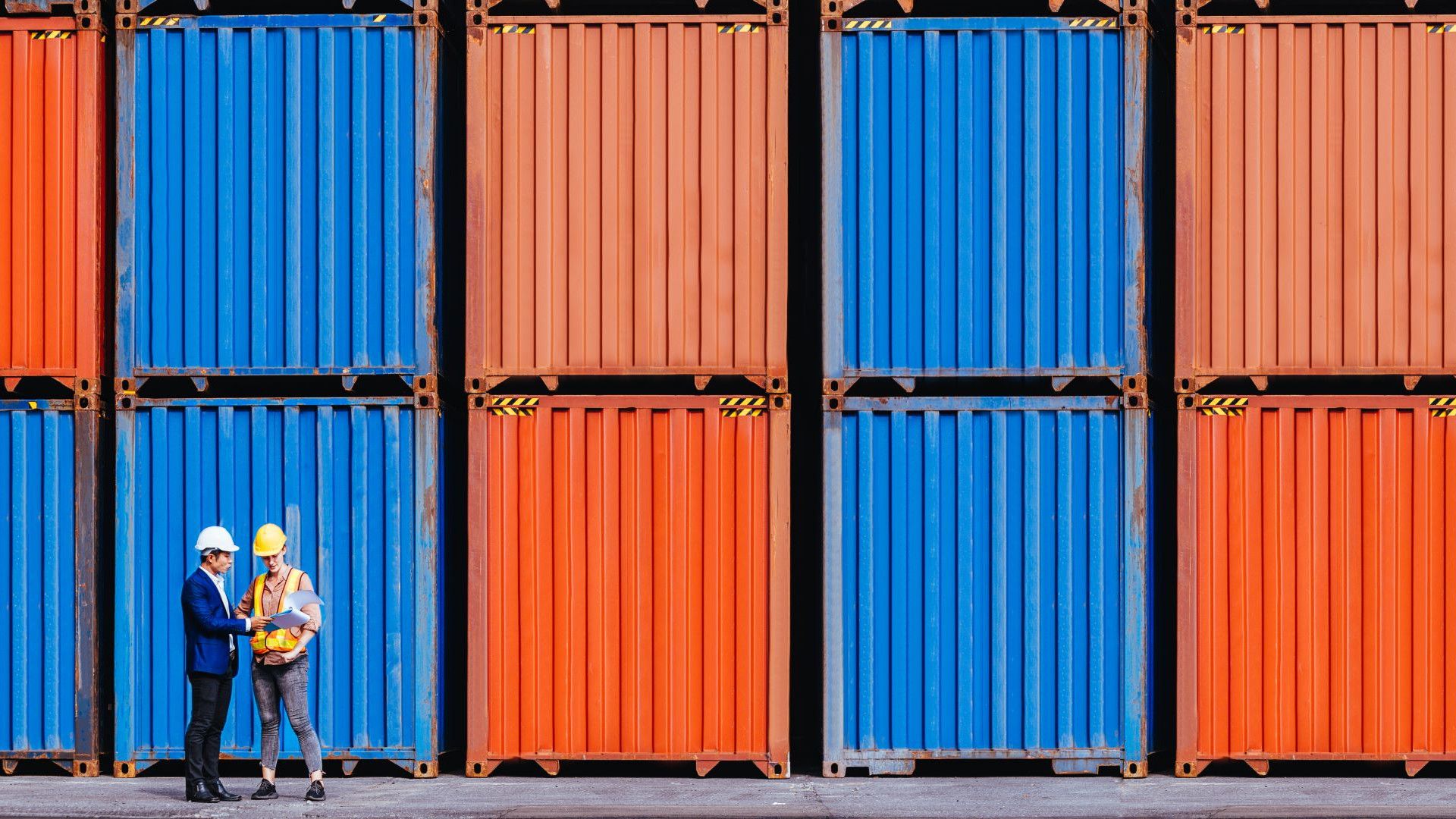 Two individuals in a shipping yard standing in front of blue and orange cargo containers. One person is wearing a white hard hat and a formal suit, and the other person is wearing a yellow hard hat, a safety vest and more casual clothing.
