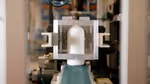 A close-up image of a white plastic bottle in an industrial testing setting. The bottle is held in place by a rectangular metallic frame.