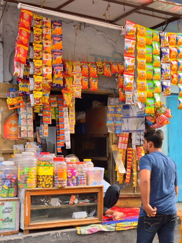 A small street-side shop displaying hanging packets of snacks and jars of candies, with a person standing in front of the shop.