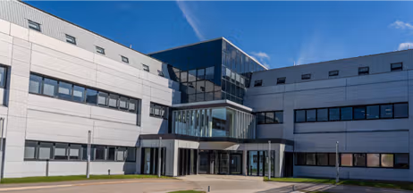 A low-level office building with a central main entrance, built predominantly from glass, with two grey wings extending to the left and right.