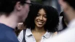 A person with curly hair wearing a light-colored collared shirt is standing outdoors among a group of people. The scene appears to be in an urban setting with blurred individuals in the foreground and background. The lighting suggests daytime, and the focus is primarily on the central figure's upper body and clothing.