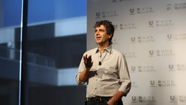 Fernando Fernandez standing on stage wearing a light-colored shirt and speaking in front of a backdrop featuring repeated “Beauty & Wellbeing” and Unilever logos, with a microphone clipped to the shirt.