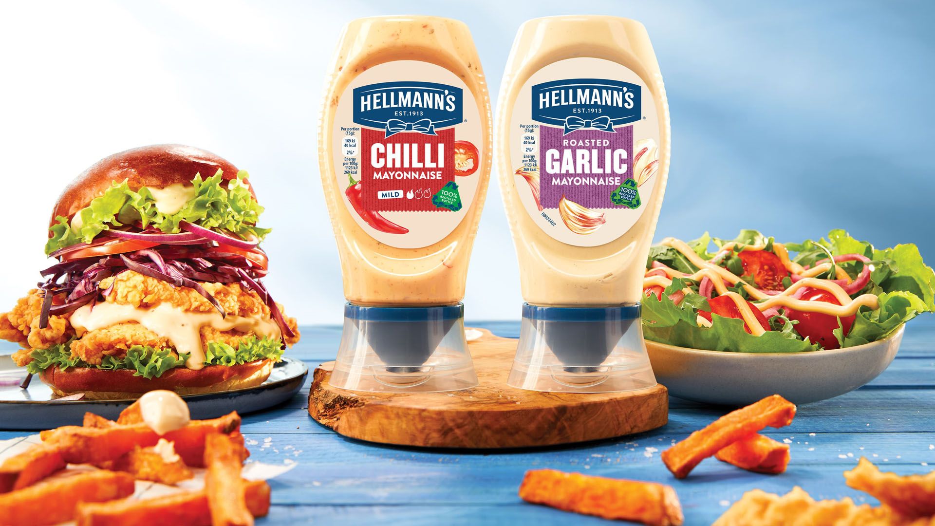 Two bottles of Hellmann’s Flavoured Mayonnaise on a table surrounded by food.