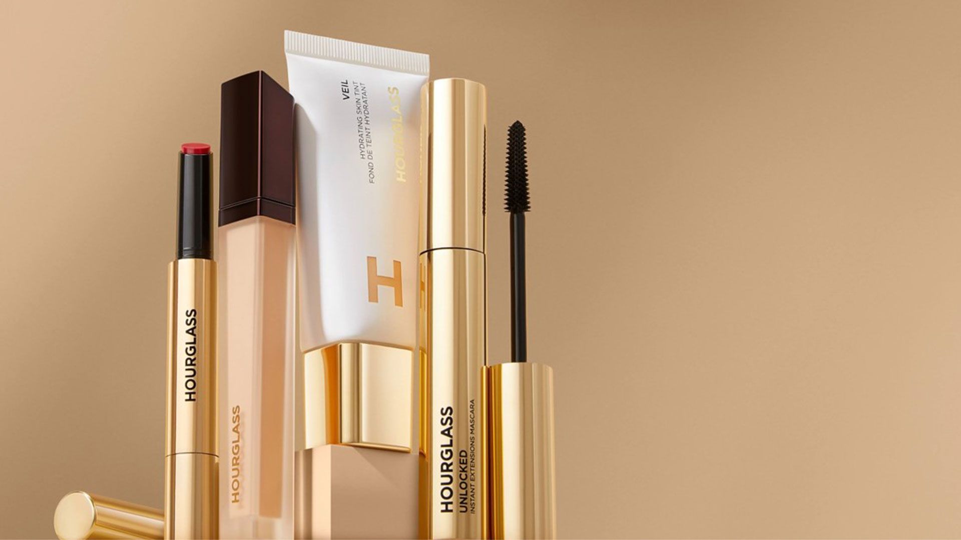 Elegant Hourglass cosmetic products displayed on a soft gold background, including lipstick, mascara, and a hydrating skin tint.