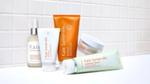A selection of Kate Somerville skincare products arranged on a white surface against a tiled background. Items include a pump bottle of Liquid ExfoliKate, a white tube of ExfoliKate Intensive Exfoliating Treatment, an orange tube of ExfoliKate Glow Moisturiser, a white jar of Exfolikate Glow, and a green-and-white tube of ExfoliKate Cleanser.