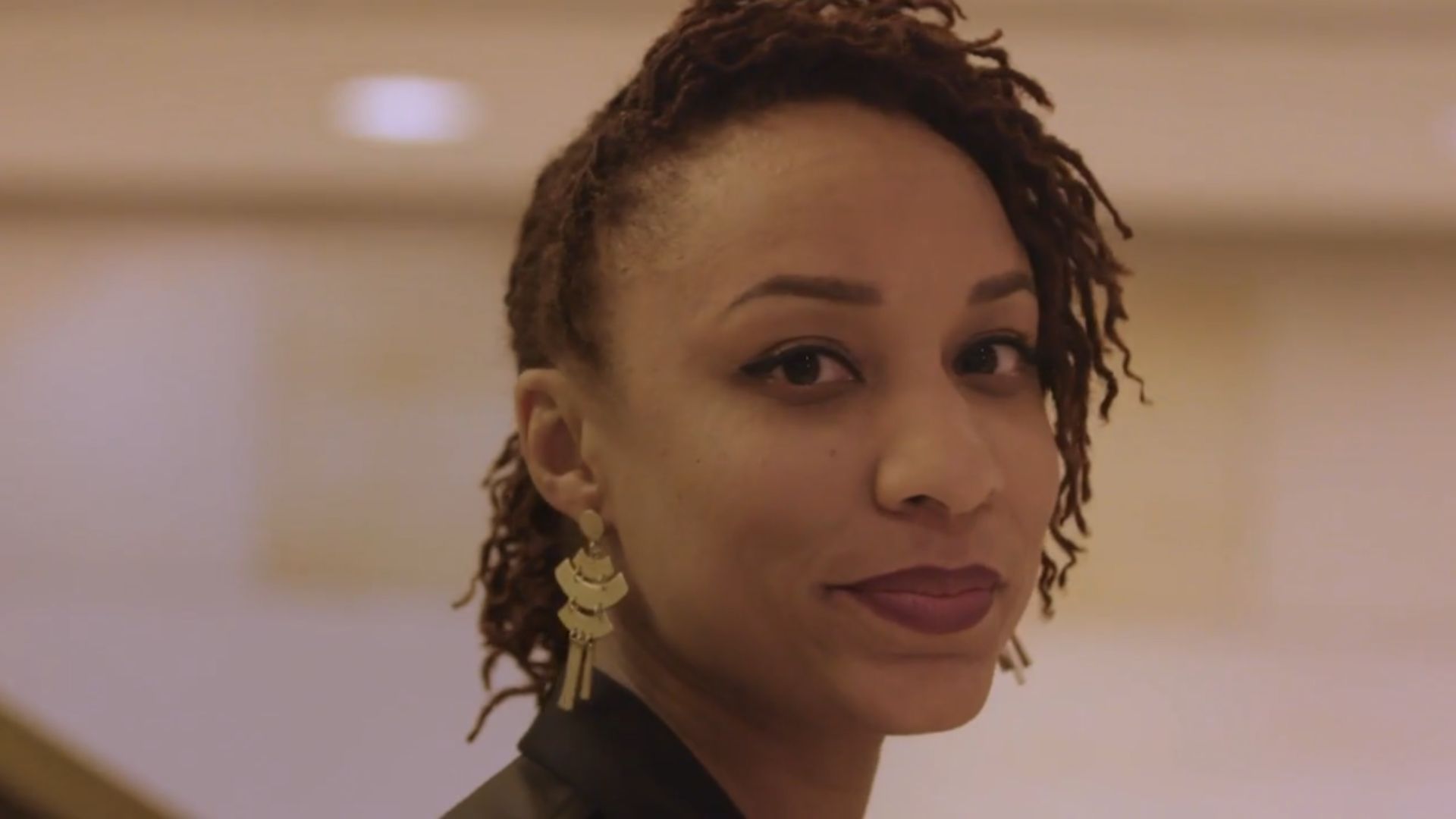 Close-up portrait of a person looking towards the camera, with short twisted hair styled to one side and a large dangling earring. The background is softly blurred with warm lighting.