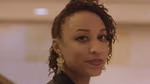 Close-up portrait of a person looking towards the camera, with short twisted hair styled to one side and a large dangling earring. The background is softly blurred with warm lighting.
