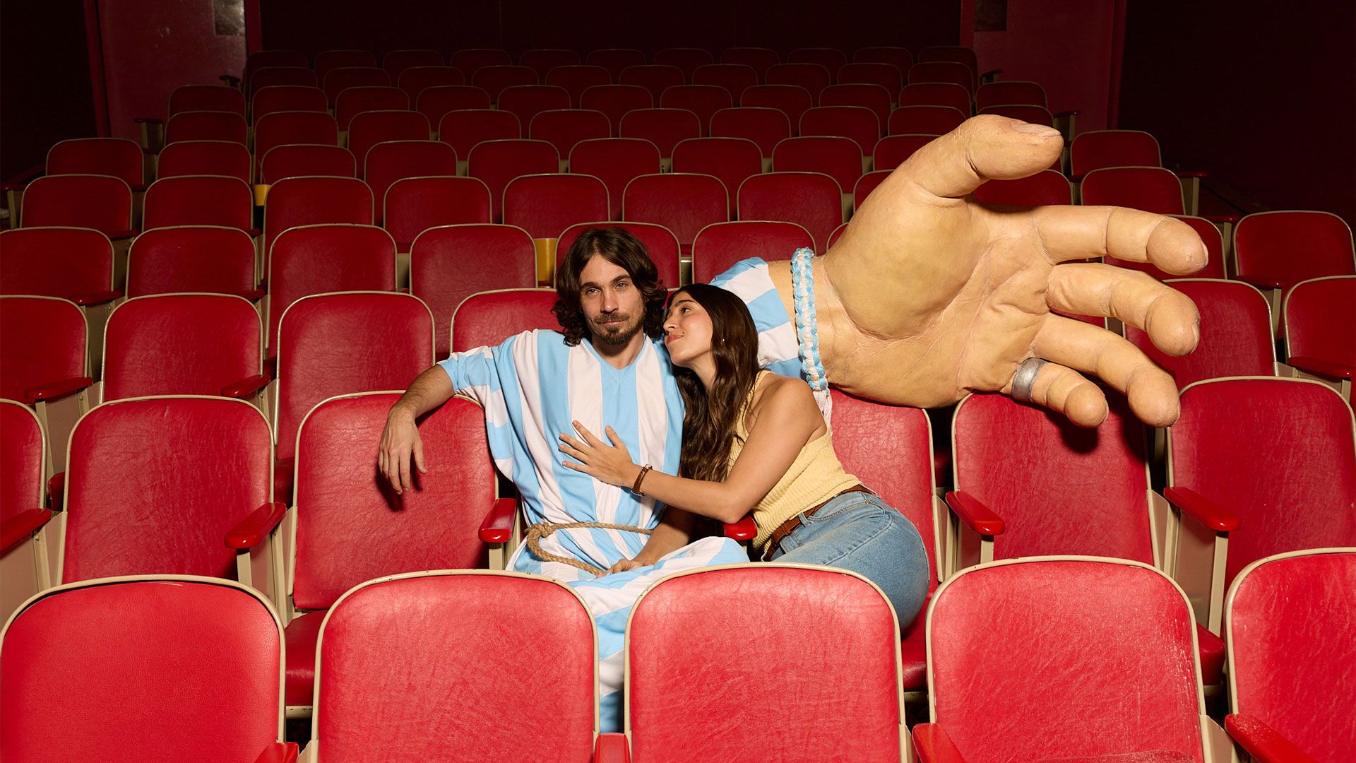 Two people sit together in a row of red cinema seats, one wearing a blue‑and‑white striped football‑style outfit, beside a large oversized hand prop resting across nearby seats.