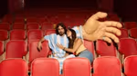 Two people sit together in a row of red cinema seats, one wearing a blue‑and‑white striped football‑style outfit, beside a large oversized hand prop resting across nearby seats.