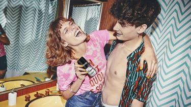 Two young people in a bathroom, with one holding a can of Axe body spray and an arm around the other's shoulder. The bathroom features a yellow sink, large wall mirror, and patterned shower curtain.