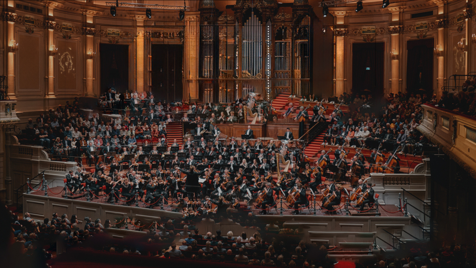 A large orchestra performing on stage in a grand concert hall filled with an audience.
