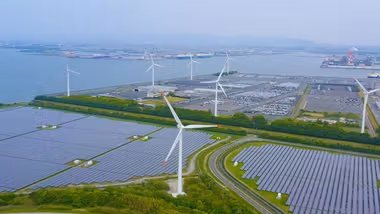 An aerial view of a renewable energy site featuring rows of solar panels in the foreground and multiple wind turbines in the background near a body of water, with an industrial port visible in the distance.