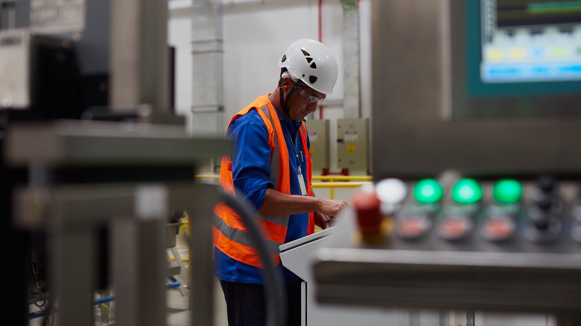 A man in a hard hat and high visibility waistcoat studies a dashboard in a factory